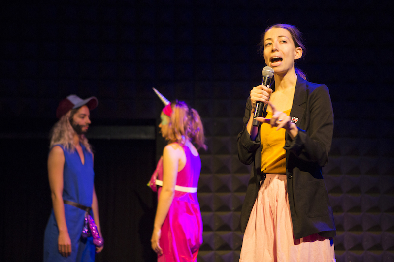 A woman in a black blazer talks to the audience while two women in brightly colored costumes are in the background. One has a drawn on brown beard.